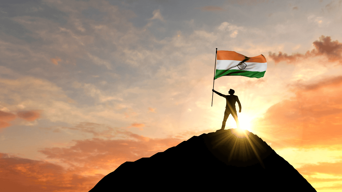 Person standing on mountain peak holding India’s national flag at sunrise, celebrating Independence Day.